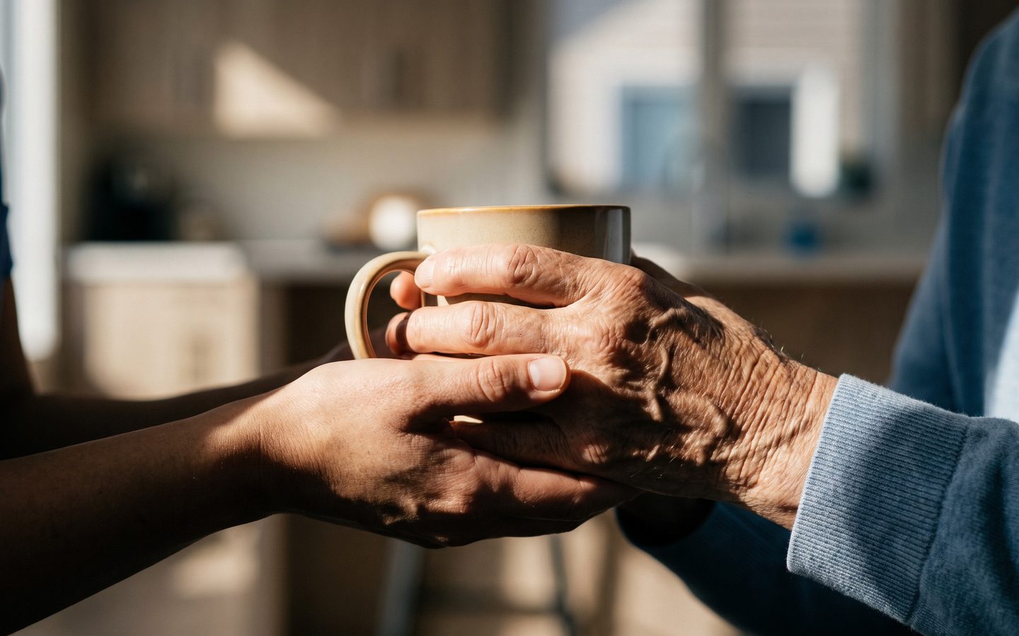 A caregiver's hands supporting an older person's hands around a warm mug