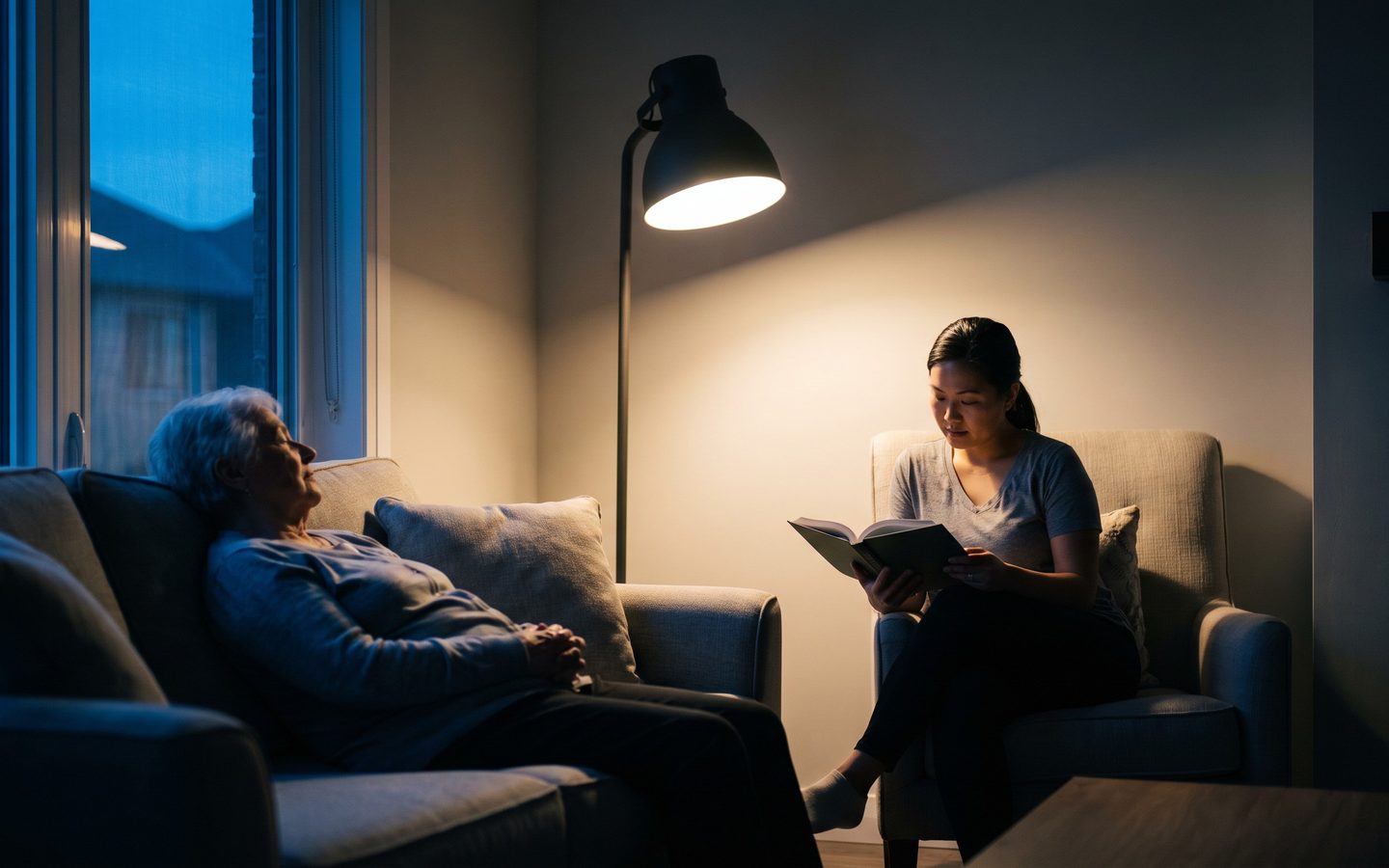 A caregiver reading by lamplight in the evening while a client rests nearby